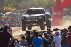 Brenthel Industries 6100 trophy truck in the 2016 Vegas to Reno race jumping over a dirt hill between a large crowd of people and a large coca cola bottle inflatable in the background