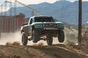 Falken Tire Brenthel industries trophy truck in the 2016 mint 400 race in Las Vegas Nevada jumping over a dirt path with a red building and mountains in the background